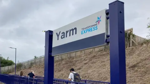 LDRS A general view of Yarm station. The image is taken from the platform and shows the sign for Yarm, with a Transpennine Express logo. It also shows a shelter and electronic signs showing the next trains. A group of people is walking up the ramp that goes from the platform to the road.