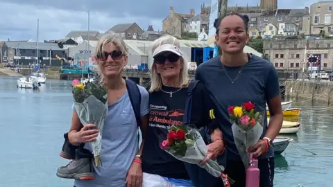 Gail Ward Three women wearing sports clothes hold a bunch of roses each and smile at the camera in front of a picturesque harbour area 