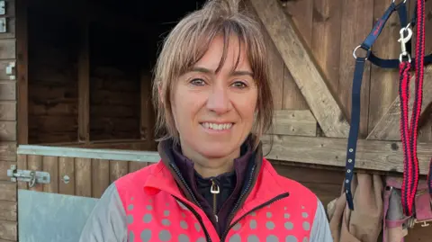 Charlotte Mitchell is smiling at the camera while standing outside a wooden horse stable. She wears a bright pink and grey reflective coat layered over a brown jumper, a purple jumper and a black zip-up jumper. She has a fringe and brown hair that has been tied up in a bobble.