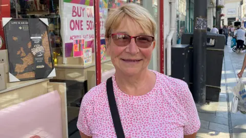 Nancy Wynn is wearing a patterned pink and white t-shirt, and a bag strap over her shoulder. She has short blonde hair, wearing sunglasses and is smiling, standing in the high street.