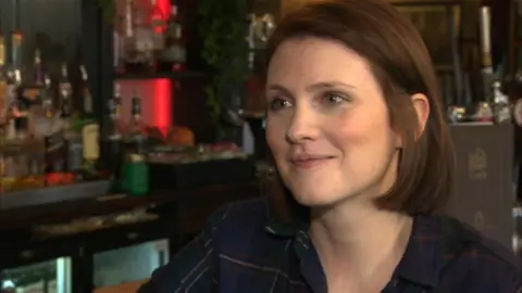 The background is a blurry area behind a bar in a pub. In the foreground is a woman in her early thirties. She is white, smiling slightly, with sharp facial features. She has brown hair in a chin-length bob, tucked behind her ears.