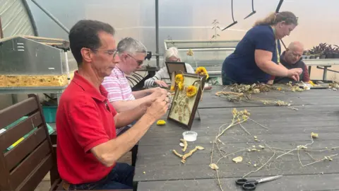 Julia Moore/BBC Four people sit or stand around a table in a greenhouse, making artwork out of flowers.