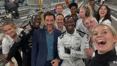 Callie Cooke/Instagram A selfie photo by Callie Cooke with her smiling alongside Damson Idris in a white motor racing suit and Javier Bardem in a blue suit. The are several other cast members in the photo all smiling and looking at the camera. They in a relatively empty pit lane at a racing circuit.