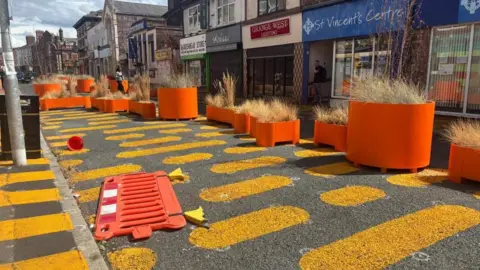 A road decorated with orange blobs painted on the tarmac. A bright orange barrier lies discarded on the carriageway. There are large orange planters along one side of the road, filled with dead grasses. There are shops along one side of the street. 