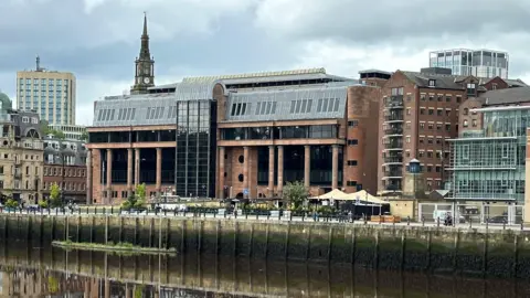 Newcastle Crown Court reflected in the River Tyne running in front of it. It is an imposing building made from smooth red stone with massive black windows and tall columns along its frontage.