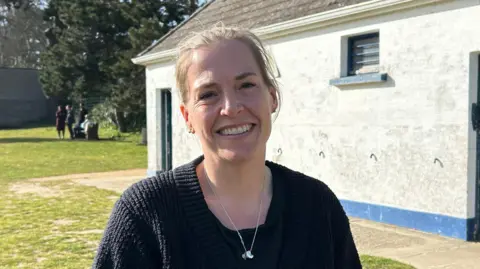 Laura has blonde hair tied back. She's wearing a black cardigan and is stood outside in a park on a sunny day. She's in front of toilet block which is white and has a blue stripe along the bottom.