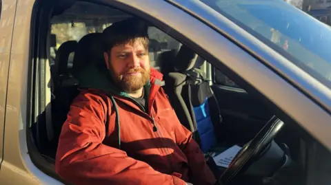 Paul Moseley/BBC Kierran Greaves sitting in the driver's seat of a hackney carriage. He is wearing a red coat over a dark top