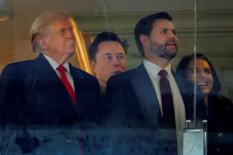 Reuters Trump wears and dark coat and a red tie, he is stood next to Vance who is also wearing a dark-coloured suit. Between their shoulders can be seen the head of Elon Musk. All three are looking to their left