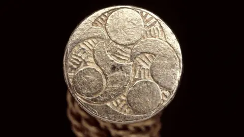 The Trustees of the British Museum A terminal of a gold Iron Age torc. It is a flat end which is decorated with a Celtic design showing three circles in a swirling design. There is a hammered pattern inbetween the circles.