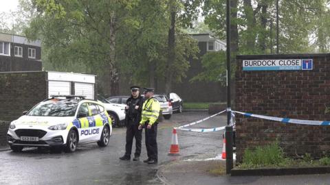 Two police officers stand in the rain beside a police car with blue and white police tape cordoning off parts of a residential area in Crawley 