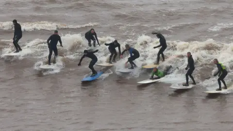Reuters Image shows a row of surfers on surfboards riding a large wave.