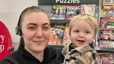 Supplied Molly Haggath is holding young Blake in her arms and smiling at the camera. They are in front of a magazine rack in the store, lined with colourful publications. Haggath is wearing a black uniform and a headpiece. She has brown hair which is pulled into a ponytail. Blake, who has short blonde hair and is wearing a brown and black jacket, has a joyful expression.
