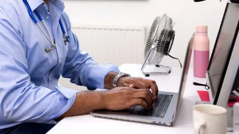 Doctor in a blue shirt with a stethescope round his neck looking at a laptop at a desk with a monitor. You can only see the body and not the head