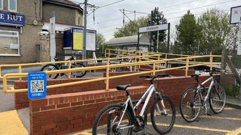 Outside Meldreth railway station. There are ramps at the entrance with yellow rails either side. Two bikes are parked at the front of the station.