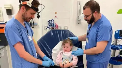 Handley Family Two doctors in blue scrubs standing by hospital bed where Peyton is lying with a Barbie doll, waiting to be examined