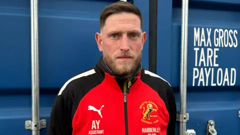 Gavin Kermack / BBC A man wearing a black and red football training top, standing in front of a blue shipping-style container. He has short dark brown hair and a brown-blonde beard.