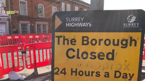 A yellow and red road sign announcing that The Borough in Farnham is closed from January to Spring sits in front of a row of red plastic road barriers around a set of roadworks.