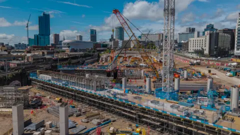 HS2 Limited A long structure is at the bottom of the photo. A yellow crane is prominent in the middle of the image and buildings are in the background.