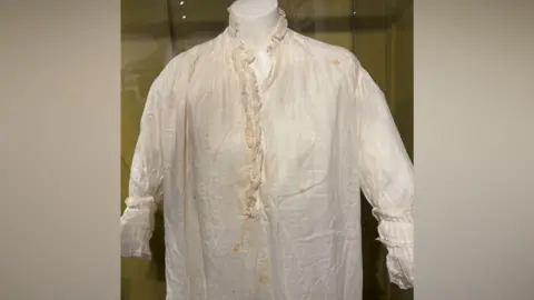 An old nightdress. It is white but has areas of yellowing. It is in a glass display case.