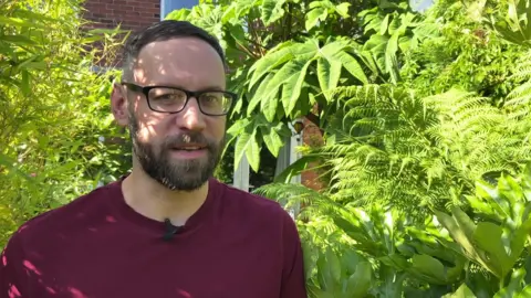 BBC/NATHAN TURVEY A picture of Dave Iveson who has dark brown hair, glasses and a beard standing in his garden. He is surrounded by lots of tropical plants and trees.