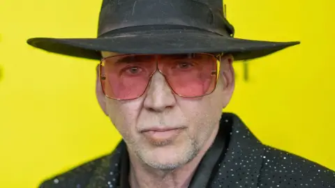 Getty Images Nicolas Cage looking at the camera in a sparkly black suit, black hat and pink sunglasses, against a bright yellow background at the premiere of The Surfer in Austin, Texas, on 10 March