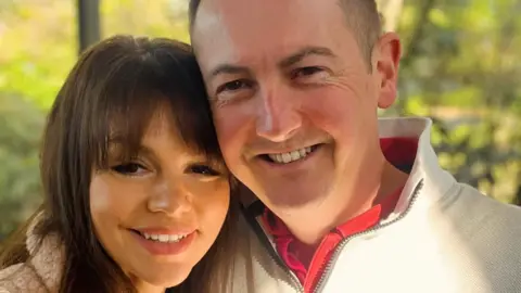 Camilla is on the left with dark hair and Matt is on the right, smiling in a white jumper. They're stood in front of greenery.