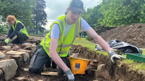 Arthur Redfern is digging in the soil. He is wearing a white t shirt, high visibility vest and cap. He also has grey gloves on. He is shovelling into a yellow bucket. In the background is another person, a grey fence and some large trees on green grass.