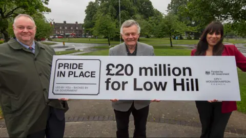A man wearing a coat, shirt and tie is on the left, another man with grey hair is wearing a grey jacket in the middle and a woman is on the right in a red jacket. They are carrying a large banner including "£20 million for Low Hill" in black letters and numbers.