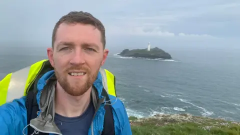 Aidan Courts wearing his hiking gear and standing by the sea on his walk so far. He is wearing a waterproof and a rucksack and there is a lighthouse on an island behind him.