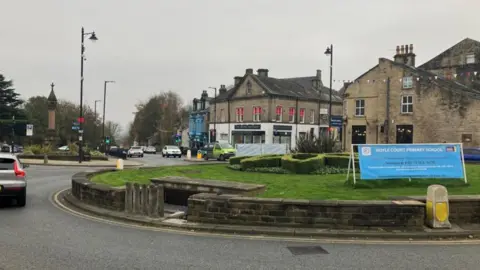 The town of Baildon in West Yorkshire. A roundabout is in the middle of a picture, with vehicles orbiting it and shop buildings in the background.