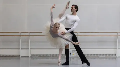 Magda Hoffman Melissa dancing with Gareth Haw in a dance rehearsal studio. She is wearing a grey leotard, white tutu, white tights and white ballet shoes. Gareth is wearing a white long sleeve top, black trousers and black socks. He has dark hair. Melissa is en pointe on her left leg with right leg in the air, she is bent forward with her arms at her side. Gareth is straight with his left leg pointed to his side. He is supporting Melissa's waist.