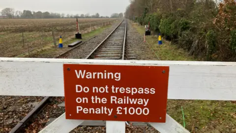 GUY CAMPBELL/BBC Red warming reading" Do not Trespass" is attached to white level crossing gates  with railway line heading off into the distance