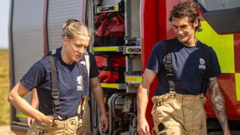 GMFRS Two firefighters stand next to a fire truck in uniform.