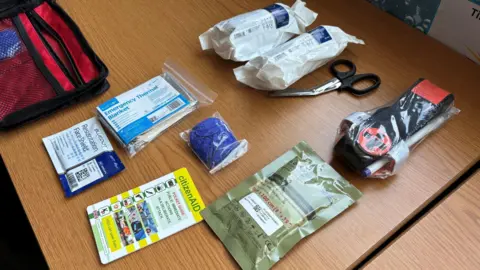 Jodie Halford/BBC Items from inside a bleed kit are laid out on a desk. This includes specially-coated bandages, a tourniquet, gloves, scissors, a foil blanket and instructions.