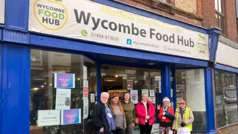A small group of people stands smiling outside the Wycombe Food Hub shop, which has a bright blue storefront and a large sign above the entrance. Posters fill the windows, and a couple of the individuals hold donation buckets, suggesting a community or fundraising effort.