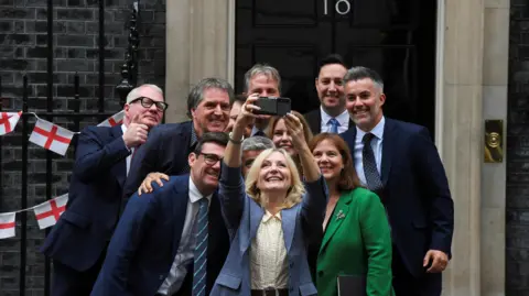 The regional mayors in England huddle together for a selfie in front of Number 10 Downing Street. A smiling Tracy Brabin holds her phone up to take the picture, with Andy Burnham to her right. There is some St George's cross bunting on the railings behind.