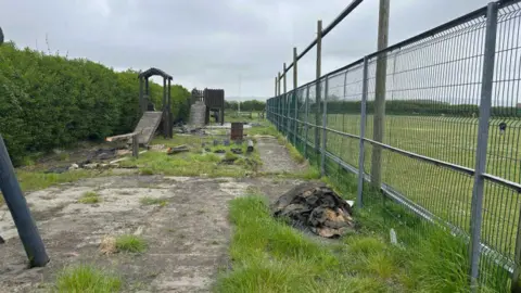 LDRS A derelict playing field area with high fences to the right of the image and abandoned wooden equipment.