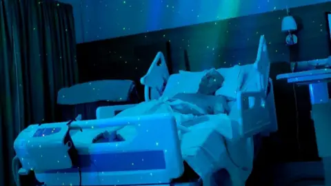Suzanne Hall A shirtless man lies in a hospital bed, propped up on a pillow, while blue and green sensory lighting surrounds him