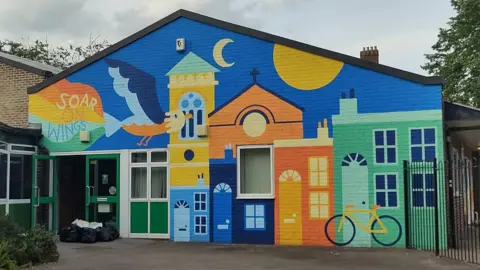 The front of the school with the multi-coloured mural. An eagle is depicted flying and trailing a rainbow with inscription "Soar on Wings".