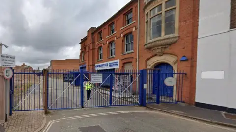 A view of 123 Northwood Street, a 20th Century brick building in Birmingham, with closed blue metal gates at the entrance and some business signs as well as a five mph speed limit. There is a blue arched door and a man in a hi-vis jacket nearby.