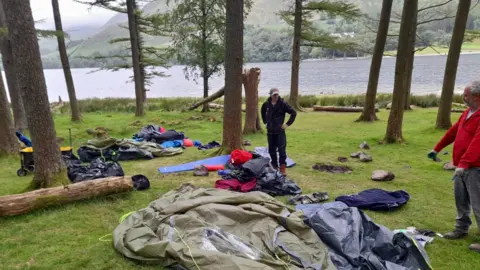 Two members of the National Trust stand at the lakeside at Buttermere with a discarded green tent in the foreground. Other items of camping equipment are strewn on the grass along with several black bin bags and items of clothing. There are several trees in the area and some have been chopped down.