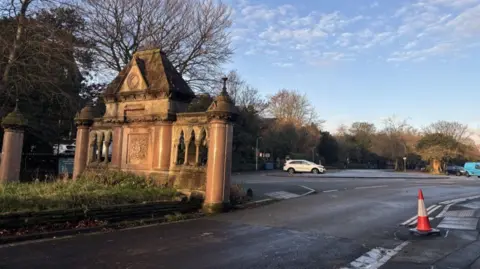 The marble gate at the road entrance to Sefton Park in Liverpool showing a traffic cone over a tarmacked area where the post was stood. Three cars are parked in the background next to the park.