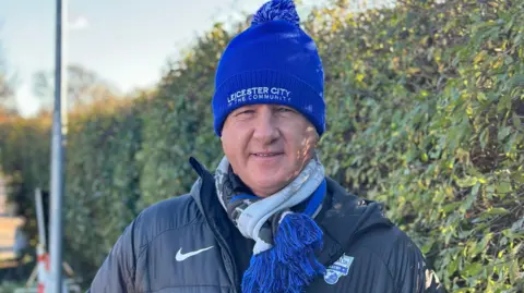 Former Leicester City player Steve Walsh wearing a hat stood next to a hedge