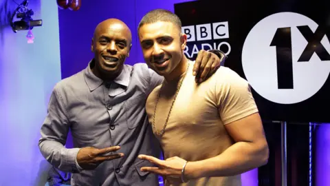 Two men stood together in a studio with purple lighting behind them. One is wearing a light grey shirt and the other a beige T‑shirt, and they are both holding up peace‑sign gestures