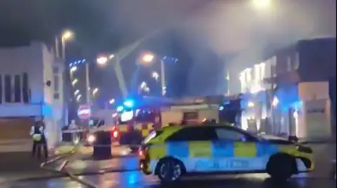 Blackpool 999s A police car is parked outside the corodned area with fire engine in background and apparatus hosing down the smoks over shops and premises at night.