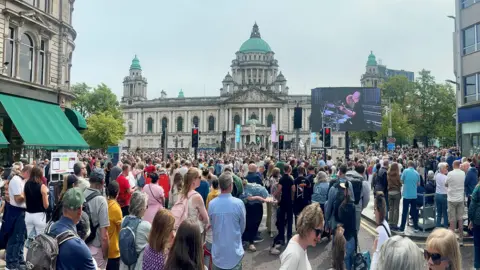 PA Media A large crowd in front of Belfast City Hall - it is a white Baroque style building with a green dome. There is a large TV screen on the right of the picture
