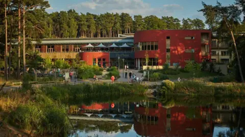 Center Parcs Center Parcs Woburn Forest: a lake edged by trees in front of a red-and-brown brick building. A reflection of the building is visible in the lake.