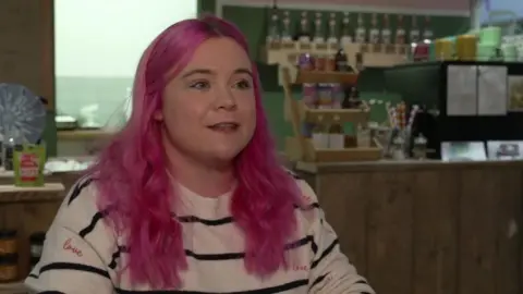 BBC A woman with long pink hair wearing a white and black striped jumper.