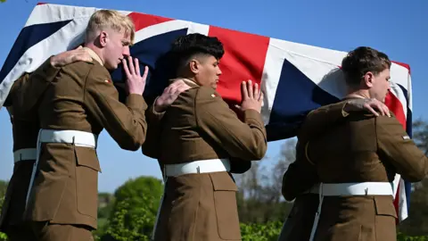 A side view of the Union Jack draped coffin holding the remains of Private Reginald Joseph Blake at the Commonwealth War Graves Commission's Loos British Cemetery Extension, on 22 April 2026. The coffin is held on the shoulders of six Royal Anglian Regiment soldiers, three of whom can be seen. Behind is blue sky and green trees. 