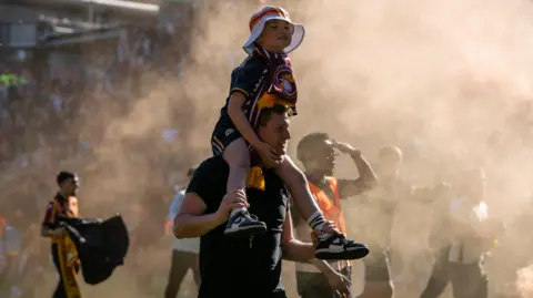 PA/Ian Hodgson A young football fan, a boy aged about six or seven, rides on his father's shoulders. He wears a claret and yellow kit and a white brimmed hat. Footballers are walking on the pitch around them and crowds can be seen in the stands in the background. The air is smoky.